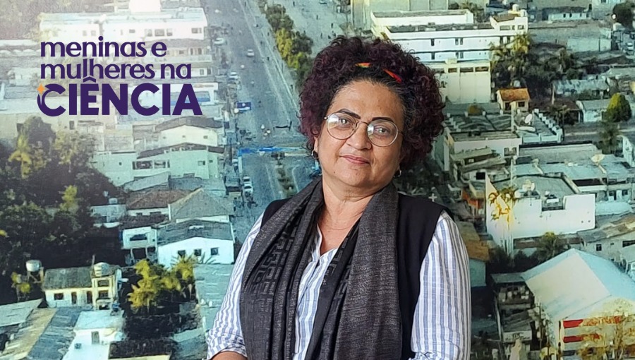 Fotografia mostra mulher negra de cabelos curtos e crespo fotografada de frente da cintura pra cima. Ela usa óculos e um tecido ao redor do pescoço. Na parte superior à esquerda, há o selo com a inscrição "Meninas e mulheres na Ciência"