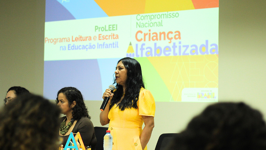 Fotografia registra a professora Sônia Regina dos Santos Teixeira falando ao microfone. Ao fundo, em uma tela é projetado a logomarca do PROGRAMA LEITURA E ESCRITA NA EDUCAÇÃO INFANTIL - PROLEEI - COMPROMISSO NACIONAL CRIANÇA ALFABETIZADA.