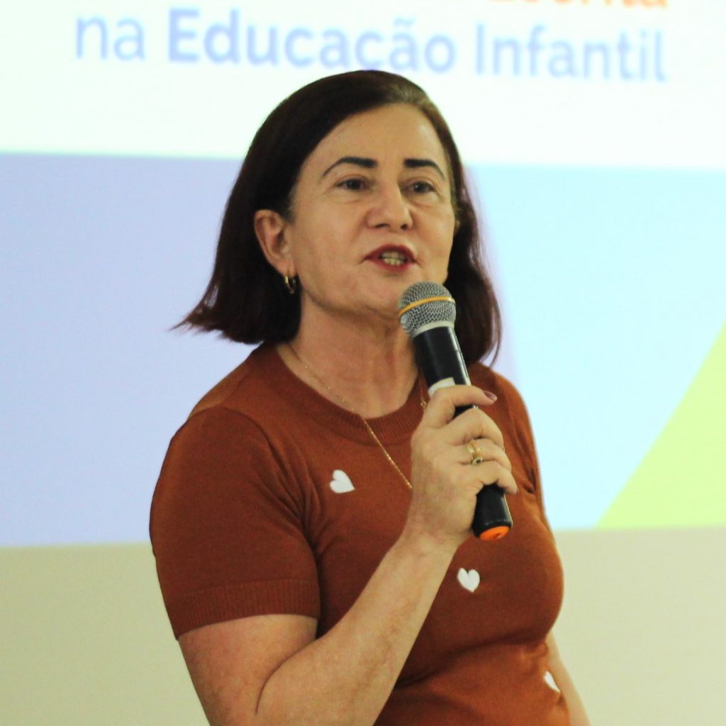 Fotografia registra a professora Celi Bahia falando al microfone. Ao fundo, é possível identificar a projeção do termo "Educação Infantil".