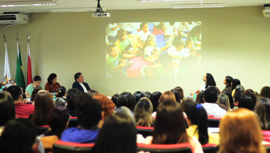 Fotografia registra parte da plateia de um auditório atenta a uma projeção feita em uma parede. Na imgem da projeção aparecem algumas crianças.
