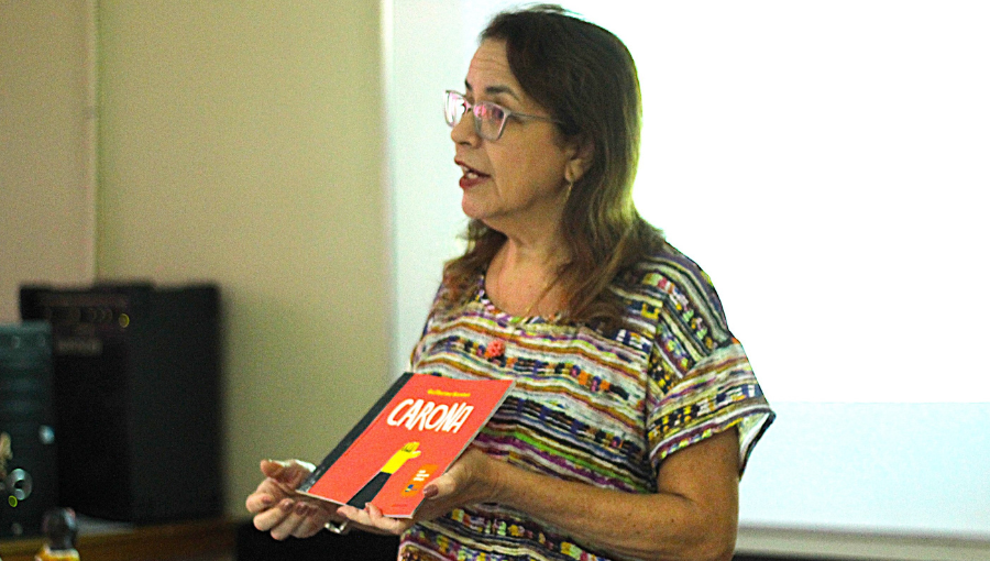 Fotografia registra uma mulher falando e mostrando um livro. o título do livro é "Carona".