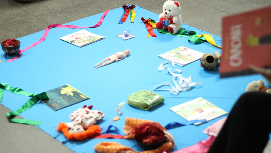 Fotografia registra diversos objetos dispostos em um tapete azul. Entre os objetos é possível identificar uma boneca de tecido e uma boneca de plástico, um urso de pelúcia, uma cerâmica marajoara, livros, fitas coloridas, entre outras coisas.