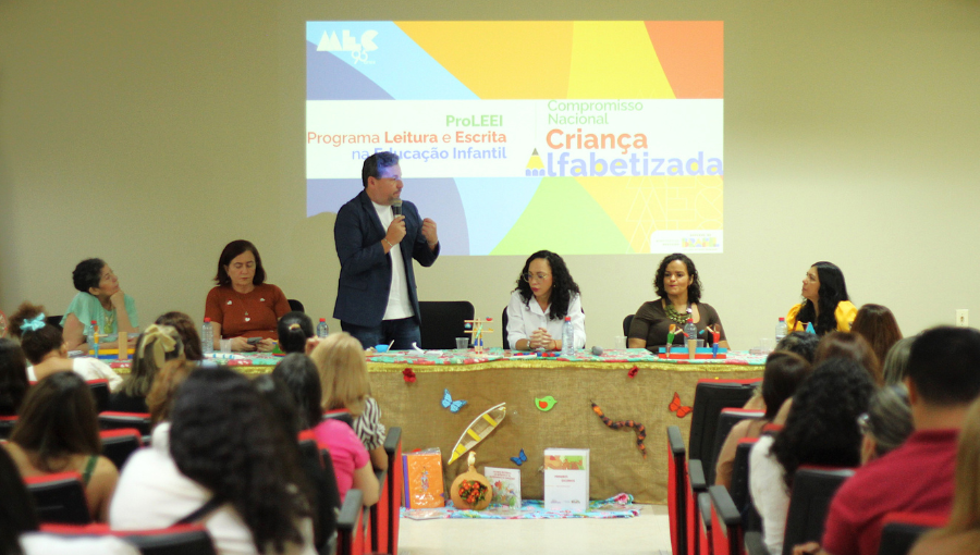 Fotografia registra a mesa de uma evento formada pelas seguintes pessoas: o Pró-Reitor de Extensão da UFPA, Nelson Souza Júnior; a coordenadora do ProLEEI CNCA-UFPA, professora Sônia Regina dos Santos Teixeira; a Diretora Adjunta do Instituto de Educação da UFPA (ICED), professora Celi Bahia; a presidente da União Nacional dos Dirigentes Municipais de Educação (Undime Pará) e Secretária de Educação de Moju, Sandra Helena Ataíde de Lima; a representante da Secretaria de Educação do Estado do Pará (Seduc), Fabiana Sena da Silva; e a articuladora estadual da Rede Nacional de Articulação de Gestão, Formação e Mobilização (Renalfa/MEC), Tatiana Cristina Vasconcelos Maia. Ao fundo, em uma tela, é projetado a logomarca do PROGRAMA LEITURA E ESCRITA NA EDUCAÇÃO INFANTIL - PROLEEI - COMPROMISSO NACIONAL CRIANÇA ALFABETIZADA.
