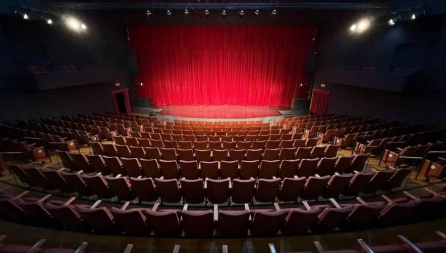 Fotografia mostra palco vazio. Há varias poltronas vazias e no palco uma ampla cortina vermelha.