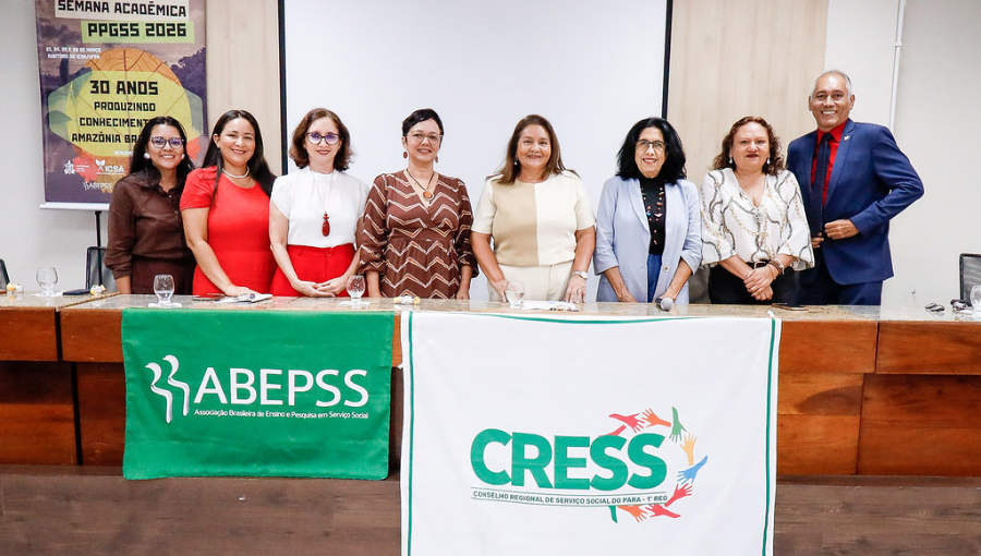 Card mostra representantes da mesa de abertura do evento, membros da coordenação do Programa de Pós-Graduação de Serviço Social, o diretor do Instituto de Ciências Sociais Aplicadas, a diretora da Faculdade de Serviço Social e representantes discentes do programa posando para fotografia atrás de uma mesa de madeira com as bandeiras da Associação Brasileira de Ensino e Pesquisa em Serviço Social e do Conselho Regional de Serviço Social.