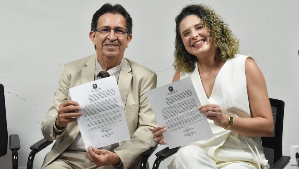 Fotografia posada de Cláudio Nery Lamarão, novo diretor-geral do IG, e Sury de Moura Monteiro, nova diretora-adjunta. Eles seguram folhas de papel e mostram para a câmera. Ambos sorriem.