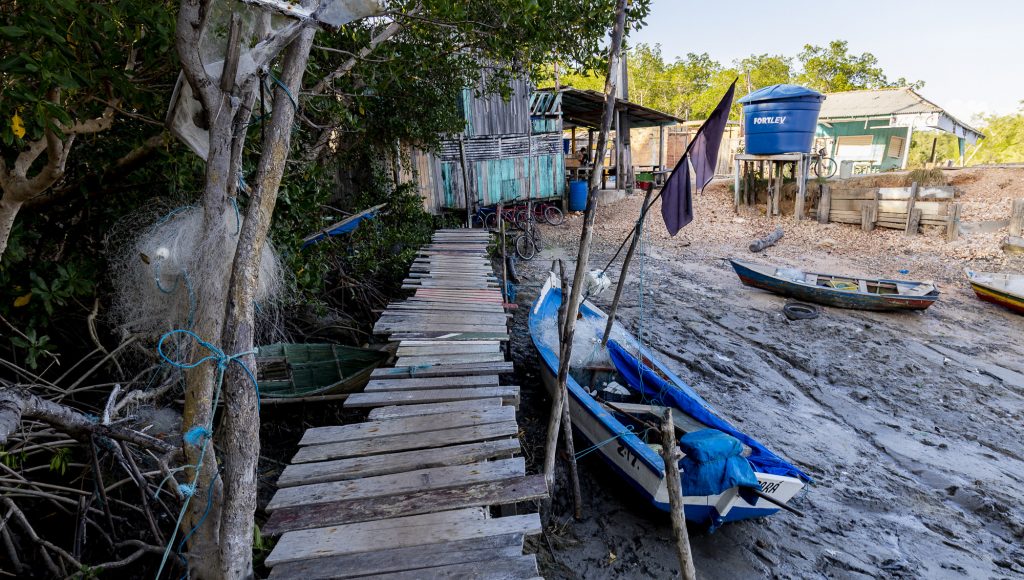 Fotografia registra a imagem de uma pequena embarcação ao lado de uma ponte de madeira. Ao fundo é possível ver casas palafitas e uma caixa d'água. O rio está seco.