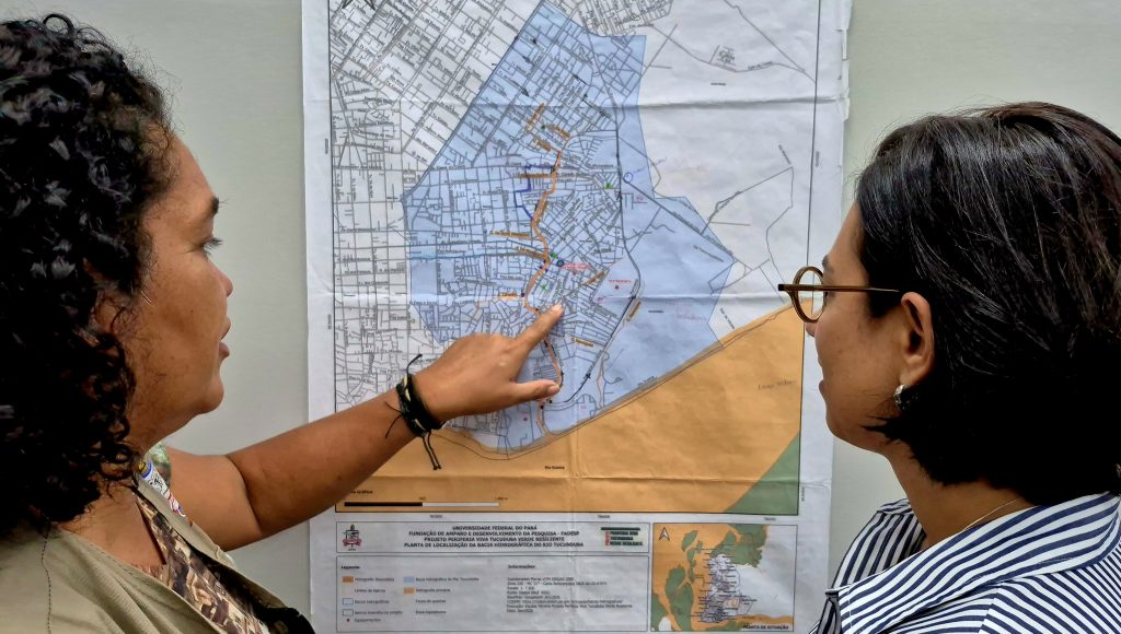 Fotografia registra duas pessoa olhando para um mapa. Uma das pessoas aponta para o mapa que está colado em uma superfície.