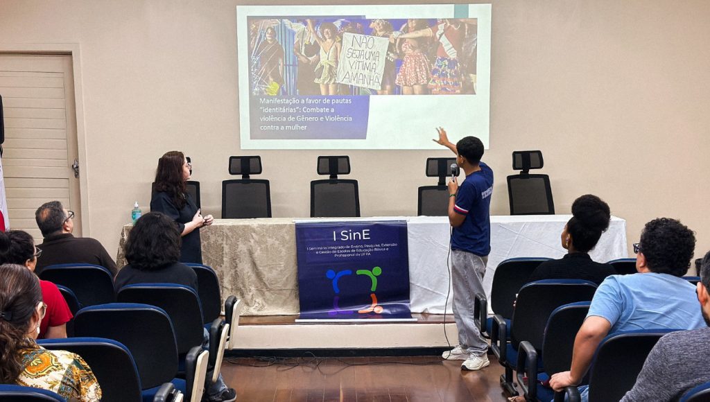 Fotografia registra parte da plateia de um auditório. Ao fundo, um jovem fala ao microfone enquanto aponta para uma imagem reproduzida na parede.