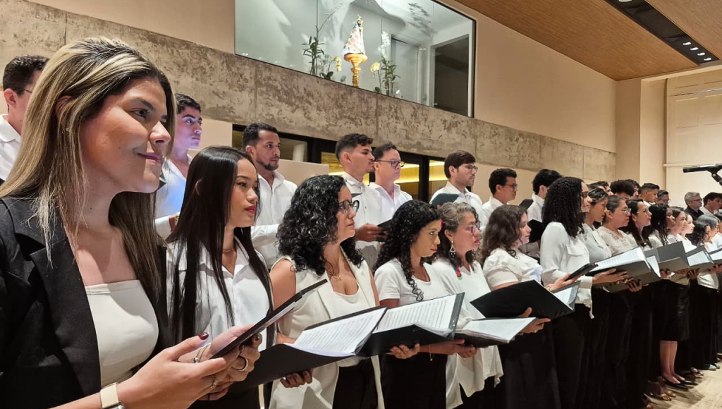 Fotografia registra um grupo de pessoas, lado a lado, com partituras nas mãos, cantando. Ao fundo, no topo da foto, é possível identificar a imagem de Nossa Senhora de Nazaré.