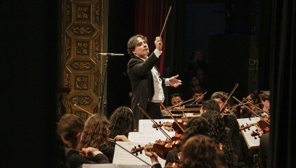 Fotografia registra um maestro regendo parte de um orquestra.