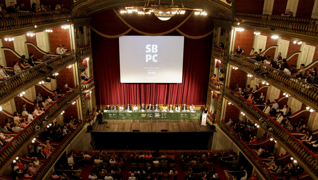 Fotografia registra a parte interna do Thheatro da Paz, em Belém. No palco, há uma grande mesa com diferentes pessoas perfiladas. Ao fundo, um enorme telão informa "SBPC". Nas laterias, é possível ver parte da estrutura dos camoretes do teztro, e no rodapé da imagem, parte da plateia que está na frente do palco.