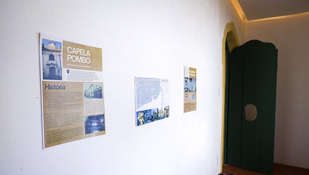 Fotografia de um ambiente interno. Ao fundo, uma porta verde com um círculo no centro. Em primeiro plano, alguns cartazes colados na parede identificam "Capela Pombo".
