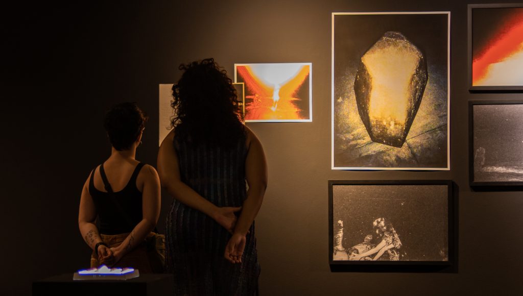 Fotografia registra duas pessoas de costa, um homem e uma mulher, observando quadros dispostos em uma parede preta.