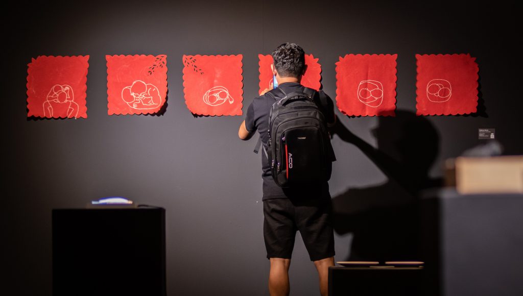 Fotografia registra uma pessoa de costa, enquanto ela fotografa quadros vermelhos dispostos em uma parede preta.