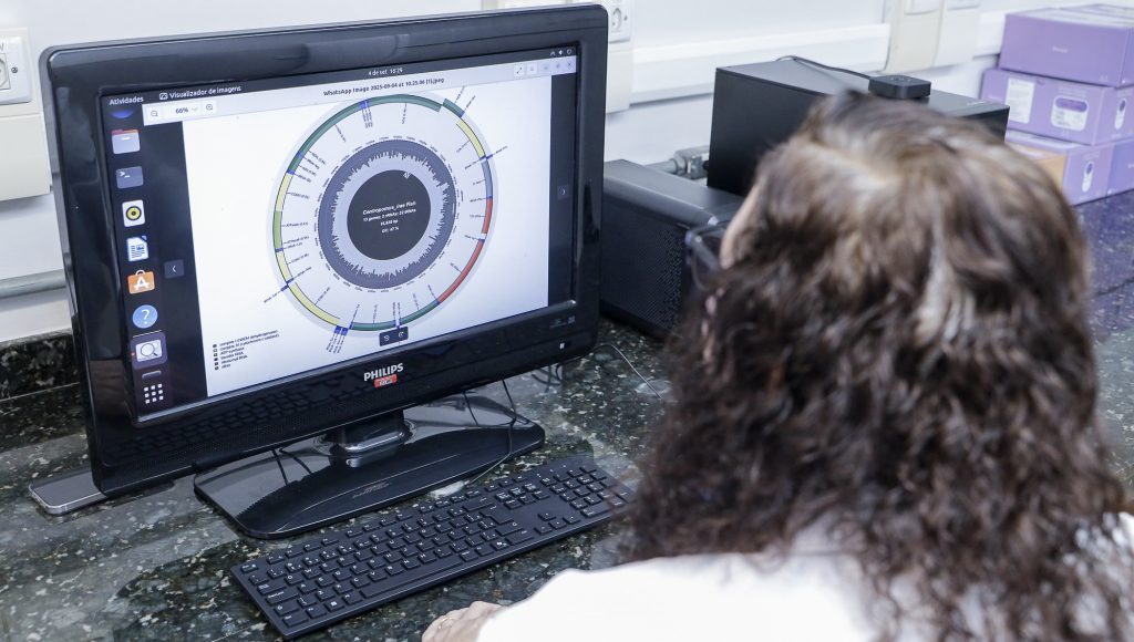 Fotografia registra uma pessoa de costa, olhando para a tela de um computador. Na tela, há uma imagem circular com termos técnicos ao redor.