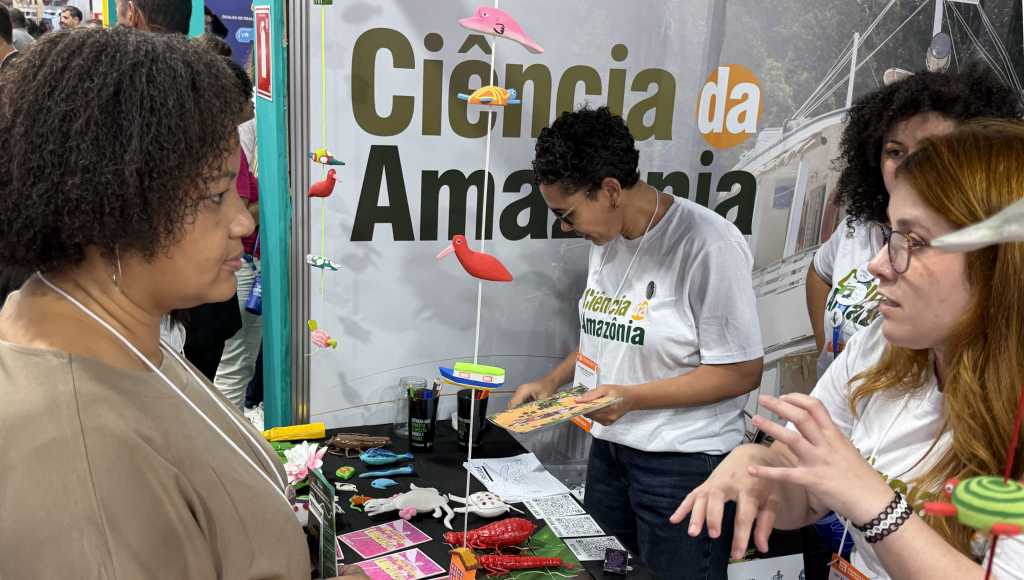 Fotografia registra uma jovem, falando e gesticulando, com uma outra mulher. Ao fundo do estande, a seguinte identificação: Ciência da Amazônia.