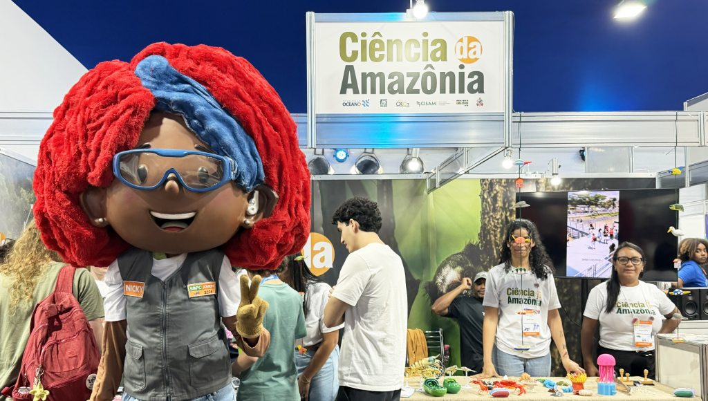 Fotografia de uma área de feira de exposições. Em destaque, no primeiro plano, uma boneca mascote negra, com cabelos ruivos e faixa azul na cabeça. Ela usa colete. Ao fundo, um estande com a seguinte identificação: Ciência da Amazônia.