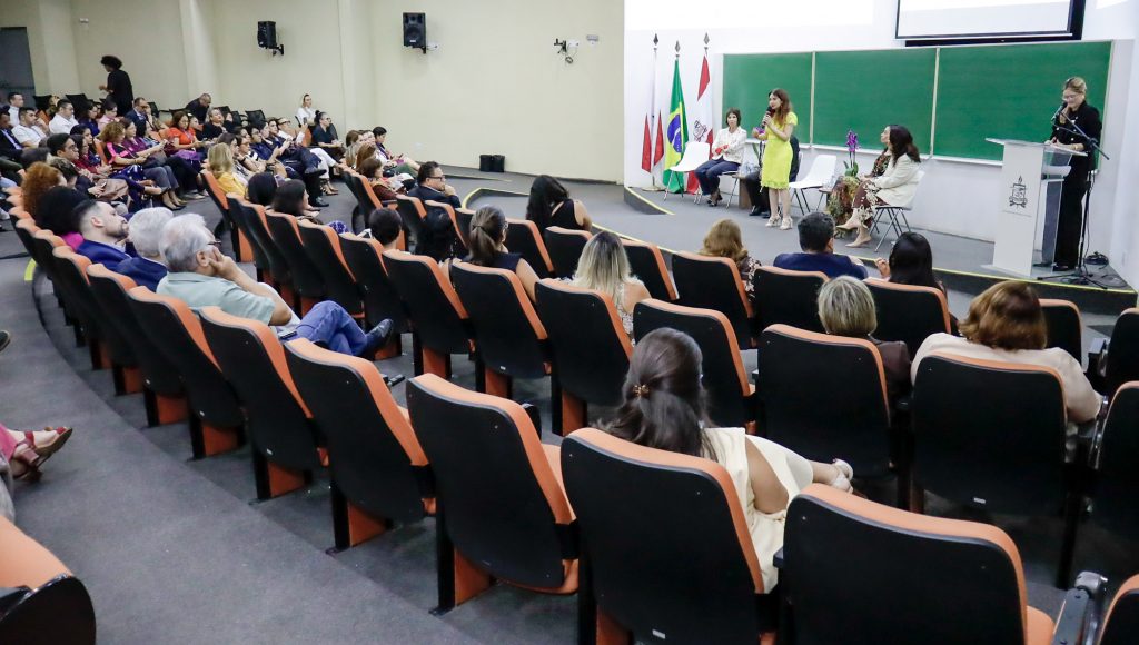 Fotografia interna de um auditório. Em primeiro plano, a plateia do auditoria sentada em cadeiras na cor laranja. Ao fundo, uma pessoa fala ao microfone.