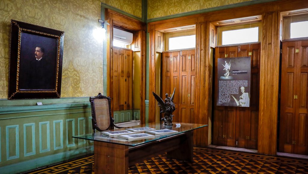 Fotografia registe para de uma sala. A sala é composta de quatro portas altas de madeira. Há uma mesa retangular de madeira com tampo de vidro onde estão disposto quatro cadernos e uma escultura. Atrás da mesa, há uma cadeira de madeira. Ao fundo, uma grande quadro com moldura de madeira. A frente de uma das portas, há também um banner pendurado com fotos e textos relacionados à escultura que está sobre a mesa.
