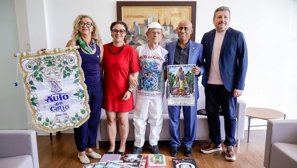 Fotografia posada de um grupo de pessoas. Entre eles estão o reitor da UFPA, Gilmar Pereira da Silva, o pró-reitor de Extensão, Nelson Souza Jr, o professor Neder Charone, a professora Inês Ribeiro e a professora Grazi Ribeiro. A professora Inês segura o estandarte do Auto do Círio e o reitor da UFPA segura o cartaz do Auto do Círio 2026.