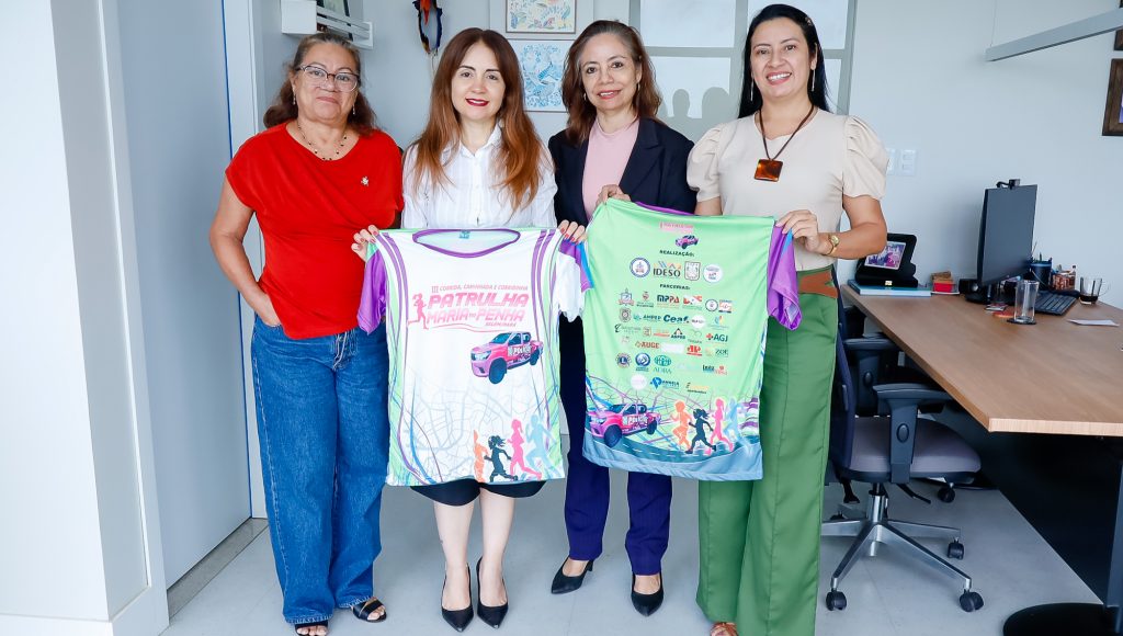 Fotografia posada de quatro mulheres. Entre elas está a vice-reitora da UFPA, Loiane Prado. A vice-reitora segura uma camiseta com a logomarca da 3ª Corrida, Caminhada e Corridinha Patrulha Maria da Penha.