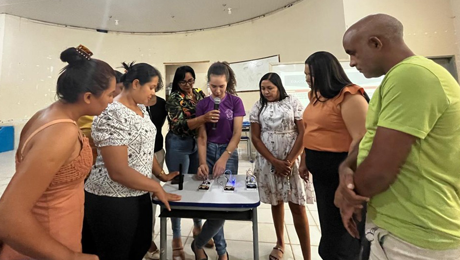 Fotografia registra uma grupo de adultos observando uma jovem manuseando objetos eletrônicos sobre uma mesa.