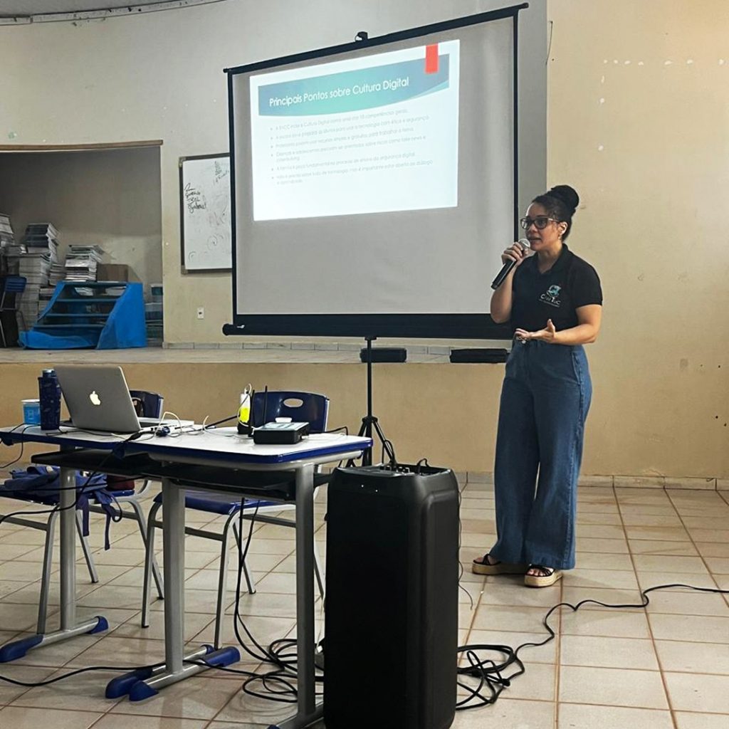 Fotografia registra a professora Viviane Almeida, falando ao microfone, a frente de um telão. No telão está sendo reproduzido um slide com a seguinte identificação: PRINCIPAIS PONTOS SOBRE CULTURA DIGITAL.