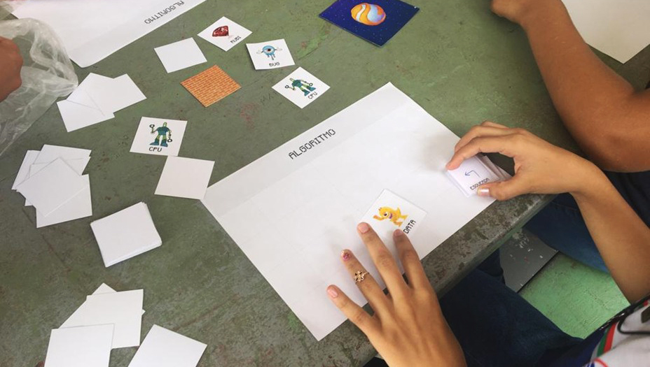 Fotografia registra duas mãos manuseando cards de papel sobre uma folha A4; A folha está identificada com a palavra "Algoritmo". Entre os cards, há cards com a palavra DATA, CPU, ESQUERDA.