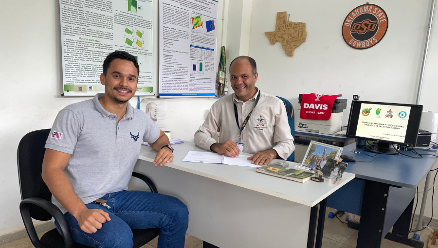 Fotografia posada de duas pessoas em um ambiente fechado. Na imagem há dois homens que estão sentados. Entre eles há uma pequena mesa. os dois sorriem. Um dos homens usa uma camisa com o brasão da UFPA.