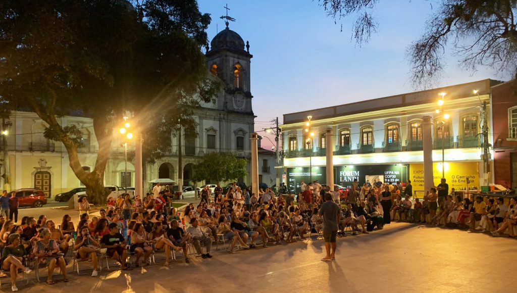 Fotografia registra uma praça com várias pessoas sentadas em um semicírculo. Ao fundo, a fachada do Fórum Landi.