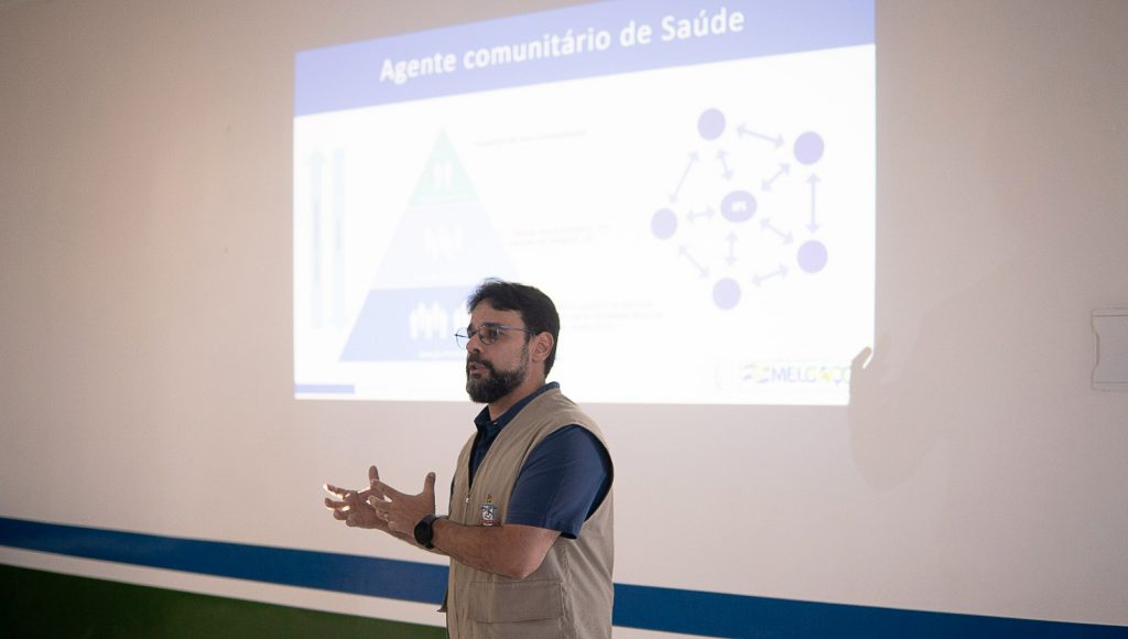Fotografia registra um homem, falando e gesticulando, em frente a um projetor que projeta um slide com a seguinte identificação: AGENTE COMUNITÁRIO DE SAÚDE.