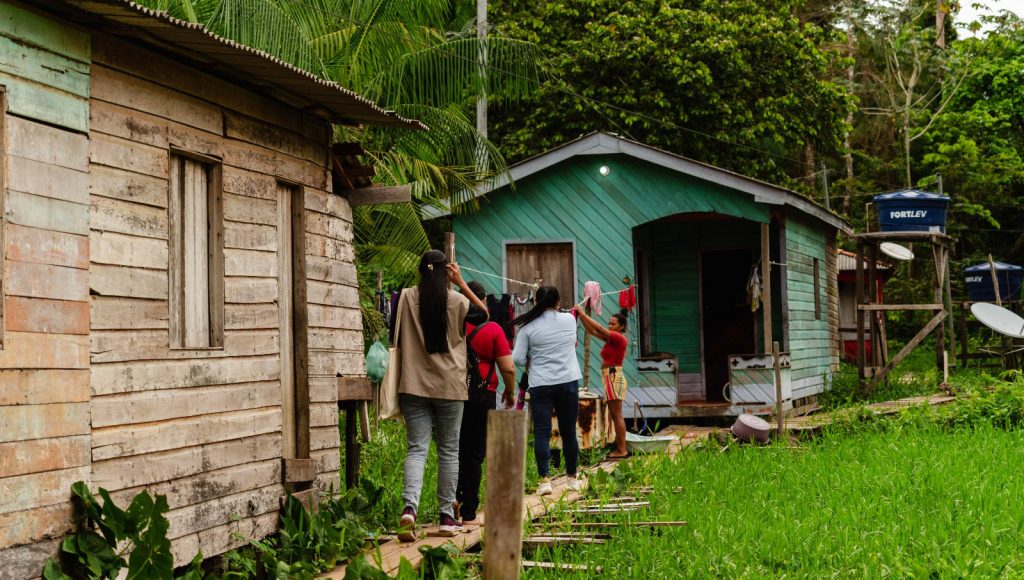 FOTOGRAFIA REGISTRA UM GRUPO DE PESSOAS CAMINHANDO POR UM caninho feito de madeira, ligando uma casa de madeira a outra. As casas e o caminho estão rodeado de mato. No fundo da imagem, é possível obsersvar caixas d´água.