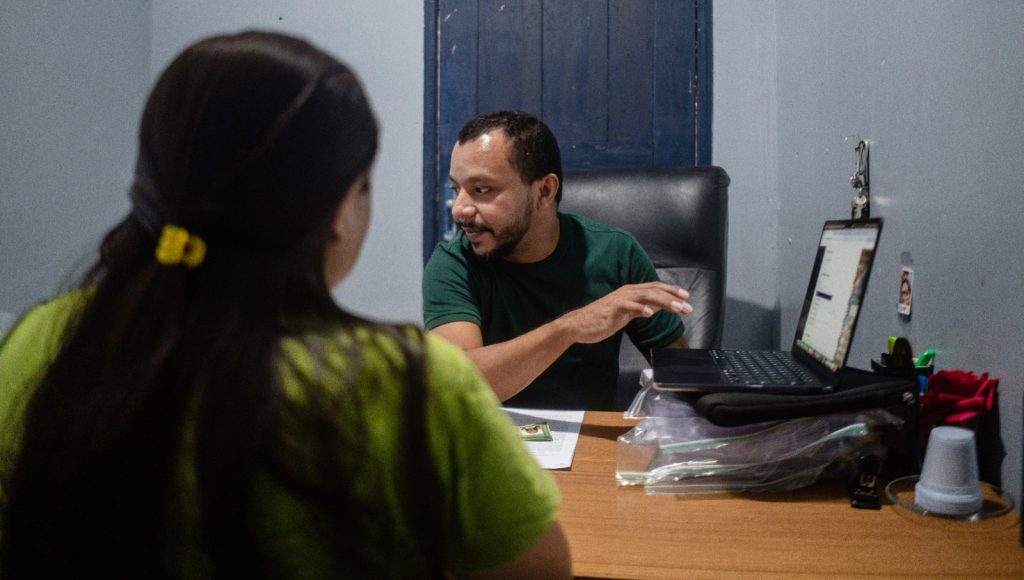 Foto registra uma homem realizando um atendimento. Ele manuseia um notebook, enquanto confere os documentos de uma mulher que está sentada a sua frente.