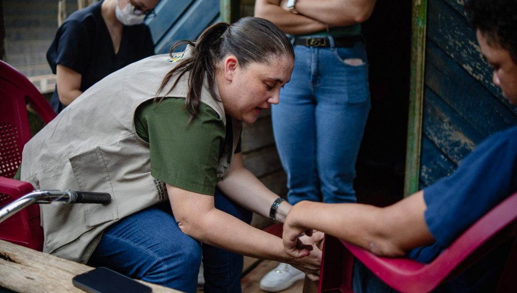 Fotografia registra uma mulher falando enquanto analisa o joelho de outra pessoa, que está sentada em uma cadeira de plástico.