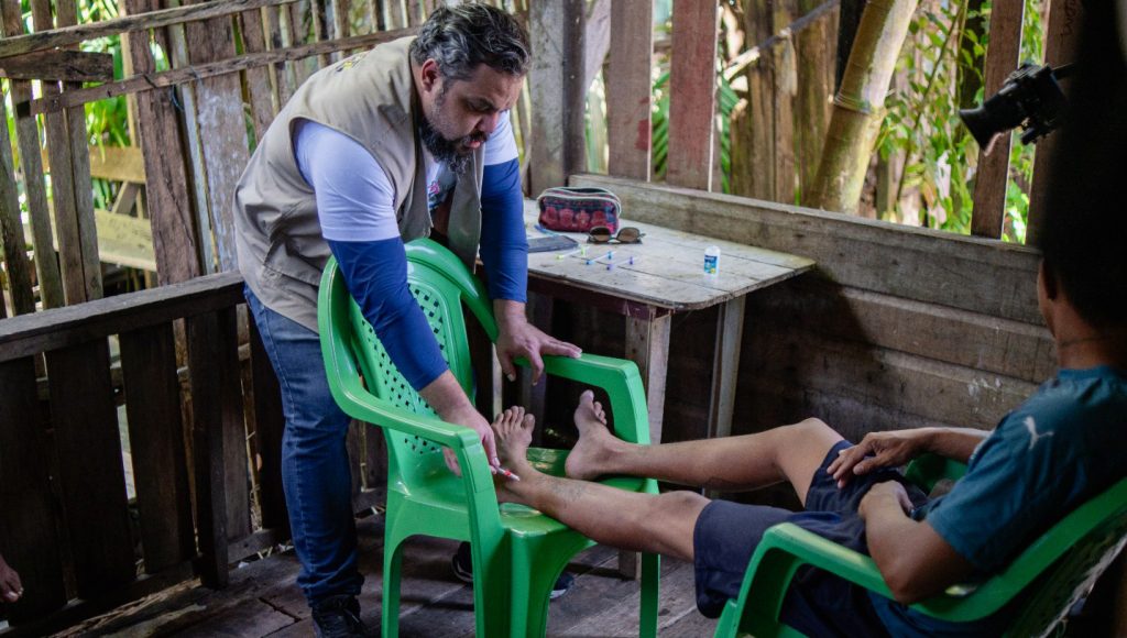 Fotografia registra um homem sentada em uma cadeira de plástico, com os pés apoiados em outra cadeira. Al lado da cadeira, um outro homem analisa a perna da pessoa que está sentada.