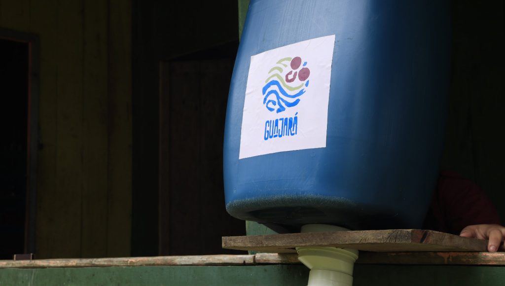 Fotografia registra parte de um recipiente azul que contem um rótulo "Guajará". O recipiente está sobre uma estrutura de madeira.