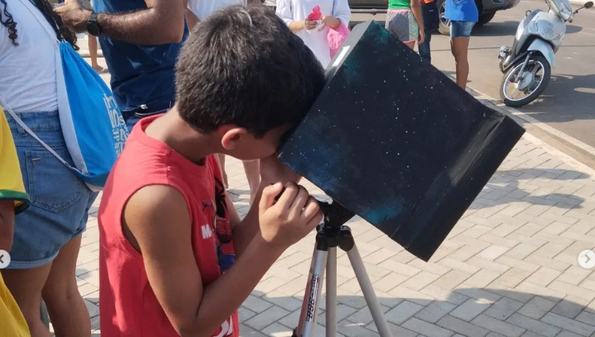 Fotografia registra uma criança manuseando uma caixa preta, sobre um tripé, em um ambiente aberto.