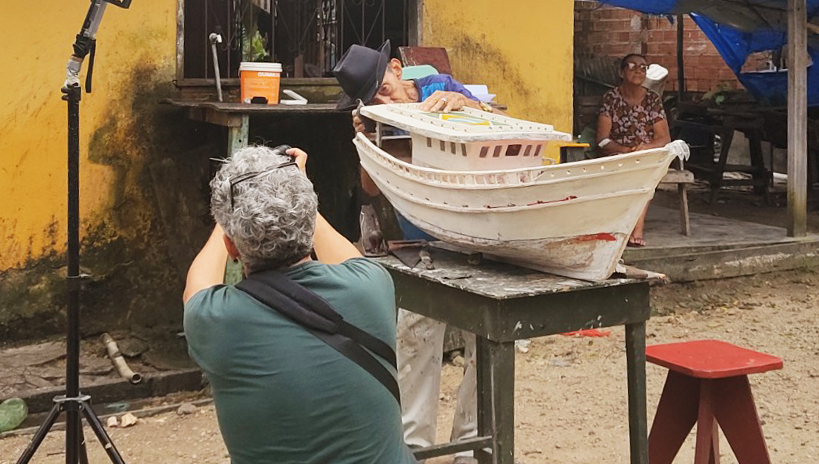 Fotografia registra uma área aberta, com chão de terra batida. em destaque na foto, um senhor manuseia uma miniatura de uma embarcação que está sobre uma mesa de madeira.De frente para o senhor, uma pessoa registra a ação com uma máquina fotográfica. Ao fundo da imagem, uma senhora observa a cena sentada em uma cadeira.