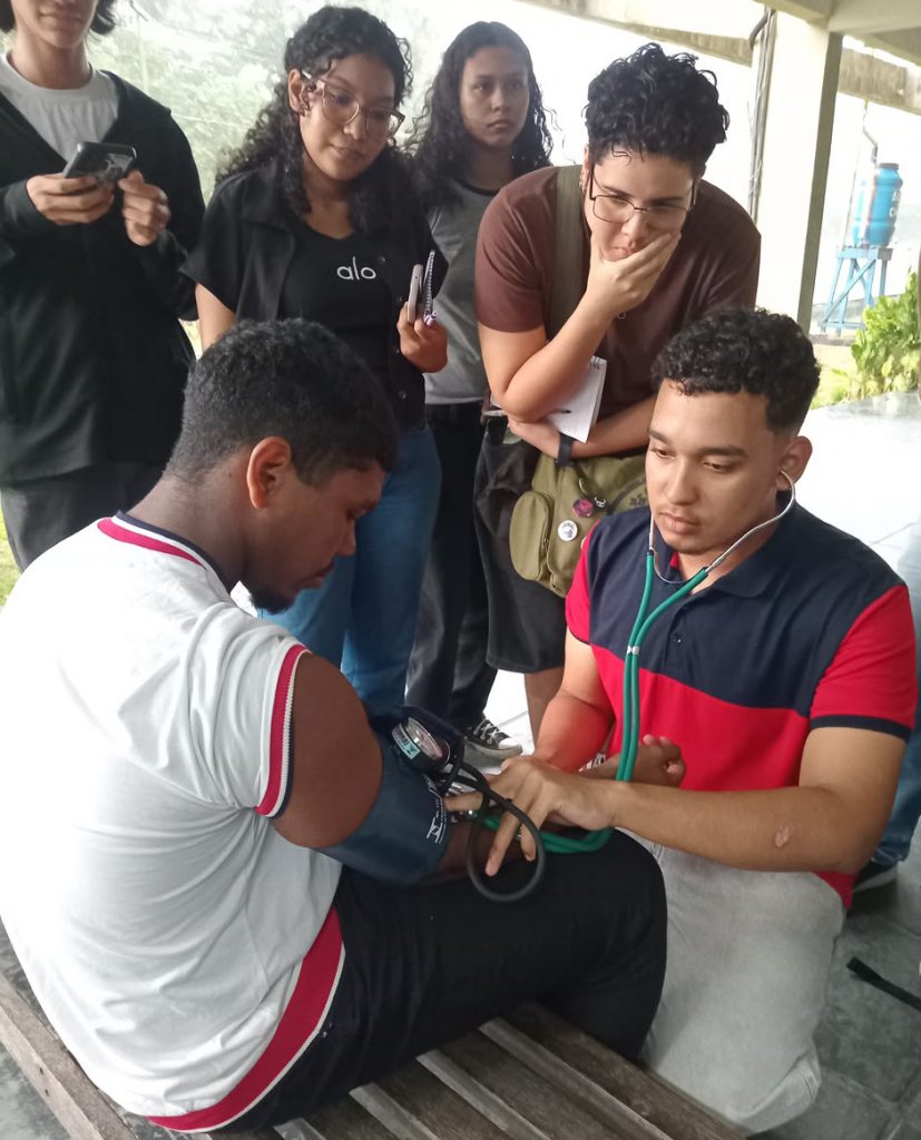 Fotografia registra um grupo de jovens atentos a um outro jovem que exercita a conferência da pressão de um outro jovens. Ele manuseia um aparelho de medir pressão e um estetoscópio.