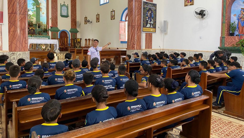Fotografia registra um homem falando para uma plateia de jovens. O homem está em pe´, enquanto os jovens estão sentados em bancos de madeira dentro de um igreja.