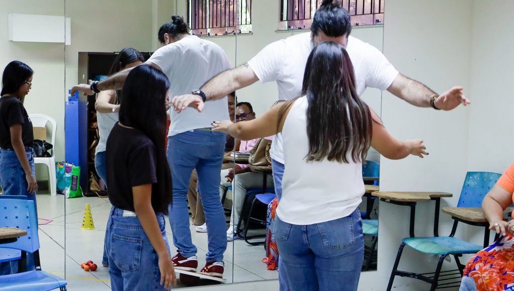 Fotografia registra três pessoas. Uma pessoa parece tentar se equilibrar sobre uma tabua circular que está sobre  um objeto que se assemelha a uma bola. Essa pessoas é observado por outras duas pessoas.