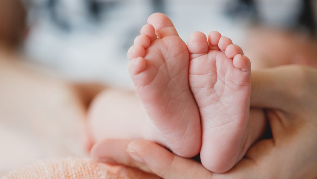 Fotografia registra uma mão segurando os dois pés de um bebê.