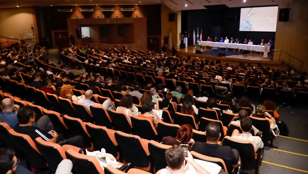 Fotografia de um grande auditório. Na foto, aparece um palco ao fundo, com uma grande mesa. A foto destaca a plateia do auditório.
