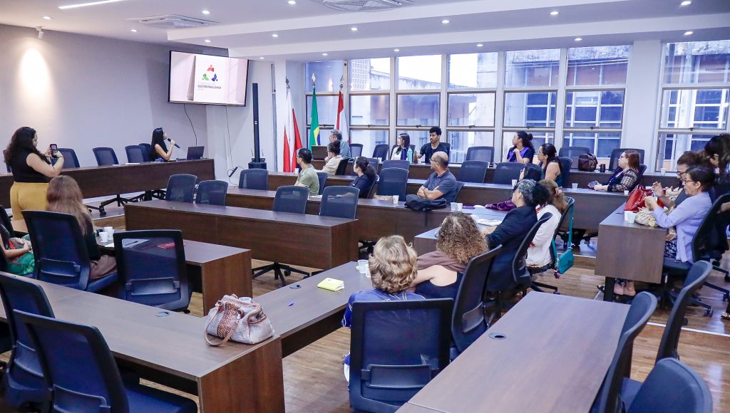 Fotografia registra uma grande sala de reuniões. Há várias mesas e cadeiras. Ao fundo, há uma tela que exibe a informação: SUSTENTABILIDADE.