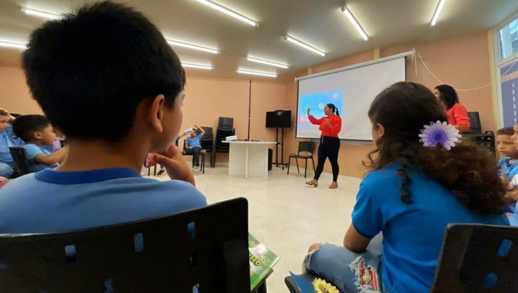 Fotografia registra uma mulher falando no meio de uma sala de aula. Ela é observada por crianças que estã sentadas em cadeiras dispostas em um círculo ao redor da professora.