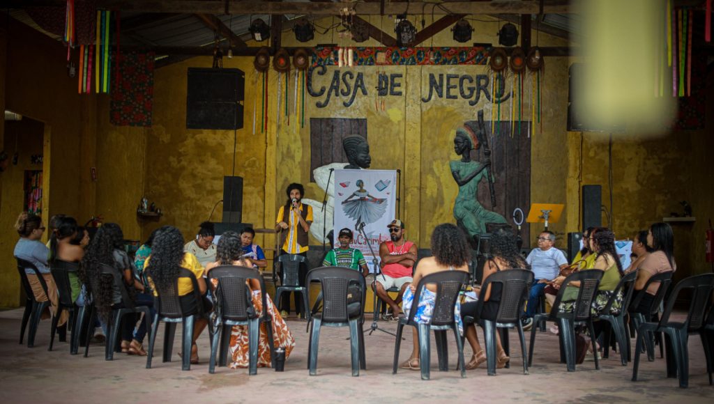 Fotografia registra um grupo de pessoas sentadas em cadeiras dispostas em um semicírculo. No centro, um homem fala ao microfone. Ao fundo, na parede, a informação: CASA DE NEGRO.