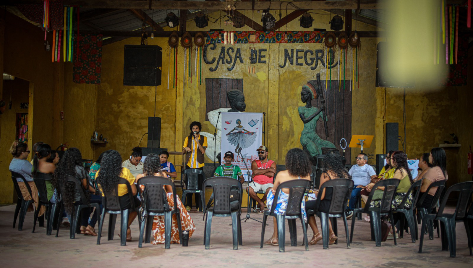 Fotografia registra um grupo de pessoas sentadas em cadeiras dispostas em um semicírculo. No centro, um homem fala ao microfone. Ao fundo, na parede, a informação: CASA DE NEGRO.