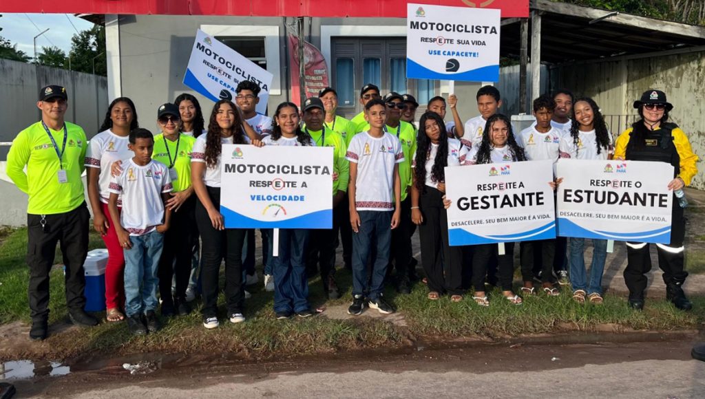 Fotografia posada de um grupo de pessoa, entre eles há jovens com uniformes escolares, e adultos com uniformes de agente de trãnsito. Alguns dos jovens seguram placas que informam: MOTOCICLISTA RESPEITE A VELOCIDADE; MOTOCICLISTA, USE CAPACETE; RESPEITE A GESTANTE; RESPEITE O ESTUDANTE.