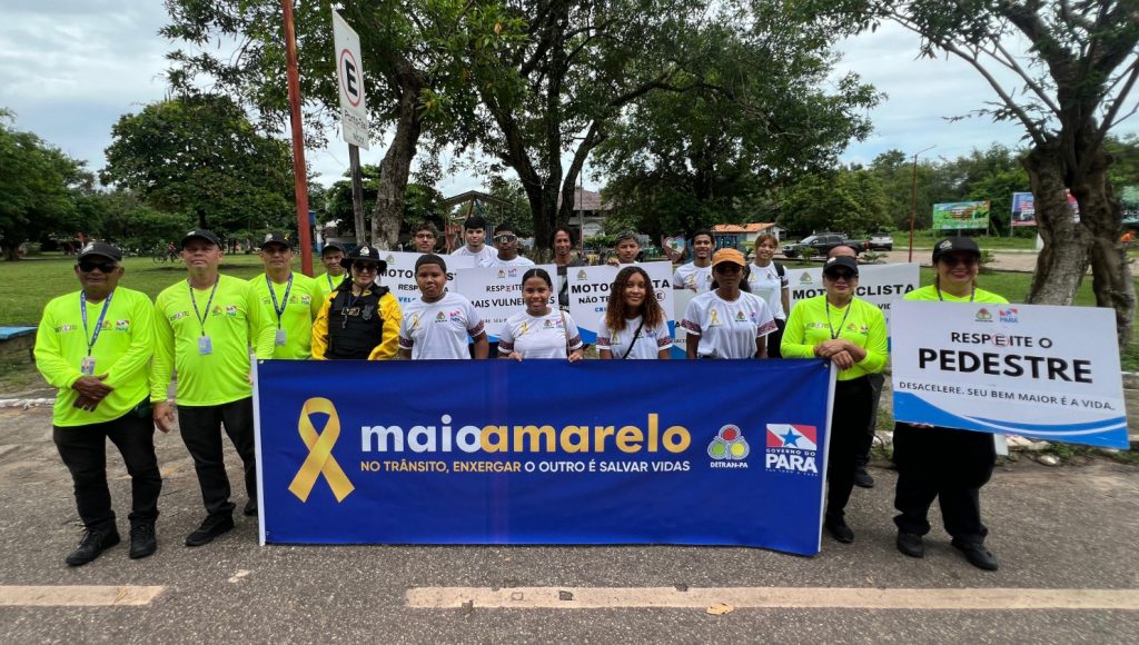Fotografia posada de um grupo de pessoa, entre eles há jovens com uniformes escolares, e adultos com uniformes de agente de trânsito. Alguns dos jovens seguram uma FAIXA que informa: MAIO AMARELO - NO TRÂNSITO, ENXERGAR O OUTRO É SALVAR VIDAS.